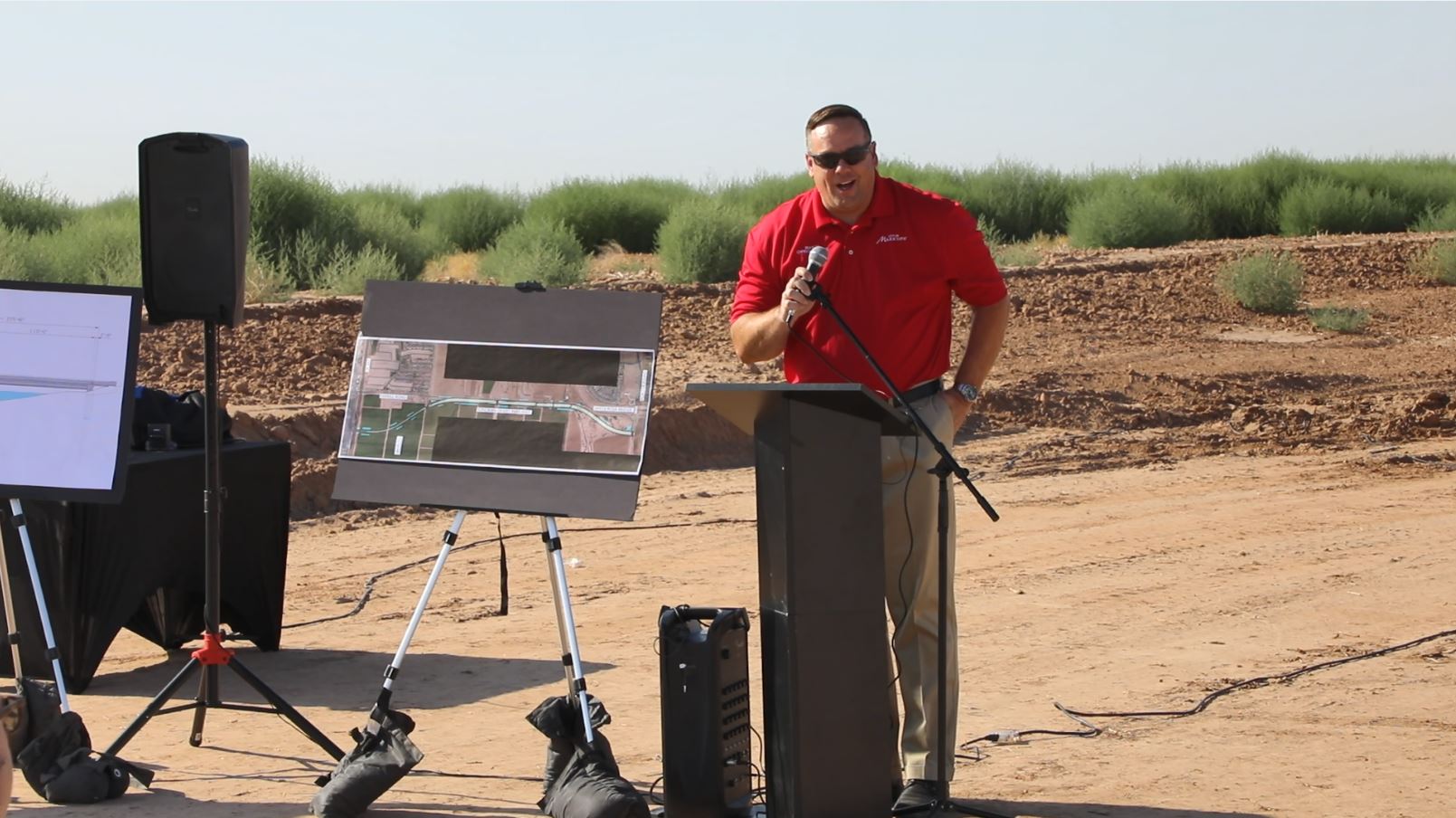 A man in a red shirt speaks at a podium outdoors on a sunny day, surrounded by desert scenery. Display boards and a speaker are set up nearby.
