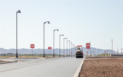 A steamroller paves a wide, empty street lined with lamp posts and 
