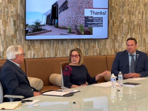 Three people in a meeting room discuss around a table with documents and water bottles. A presentation screen on the wall shows a 