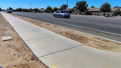 Wide view of a suburban road with a smooth concrete sidewalk on the left, cars driving on the right. Clear blue sky and houses with trees in the background.