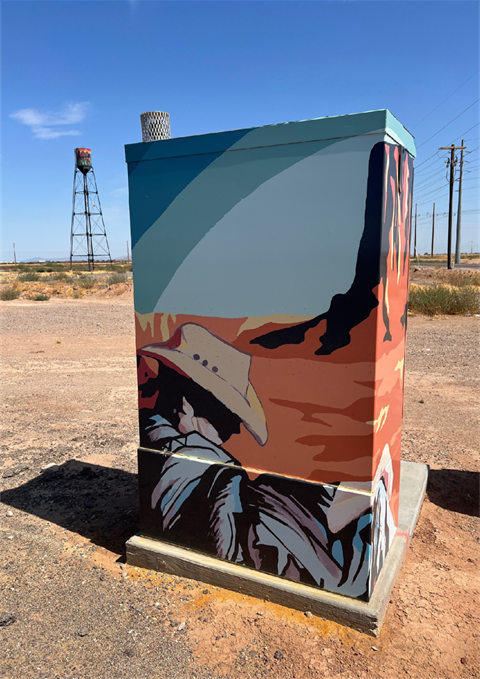 Utility box with vibrant mural of a cowboy in a desert landscape, soft hues of blue and orange. A distant water tower and sky complete the scene.