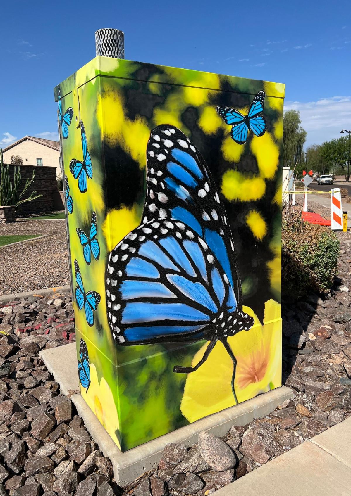 Utility box mural featuring vibrant desert scene with cactus, poppies, and mountains. A sun rises in the background. Tone is colorful and lively.