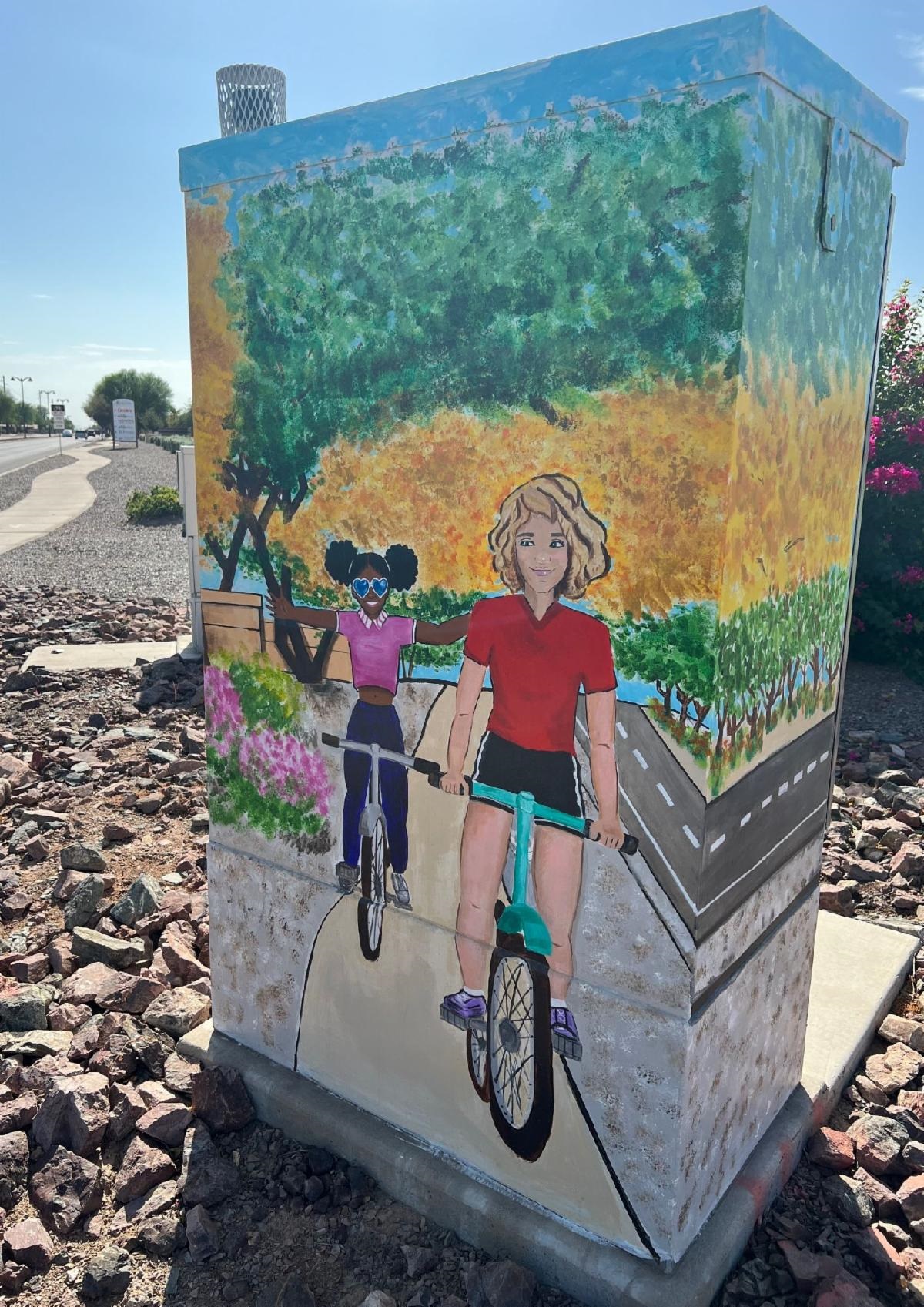 A painted utility box features two smiling children riding bicycles on a path. The background is vibrant with lush greenery and colorful flowers.
