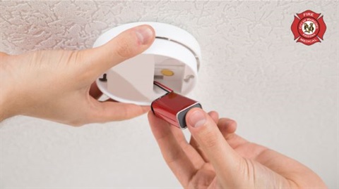 Hands replacing a battery in a ceiling-mounted smoke detector, highlighting fire safety. A fire rescue emblem is visible on the upper right.