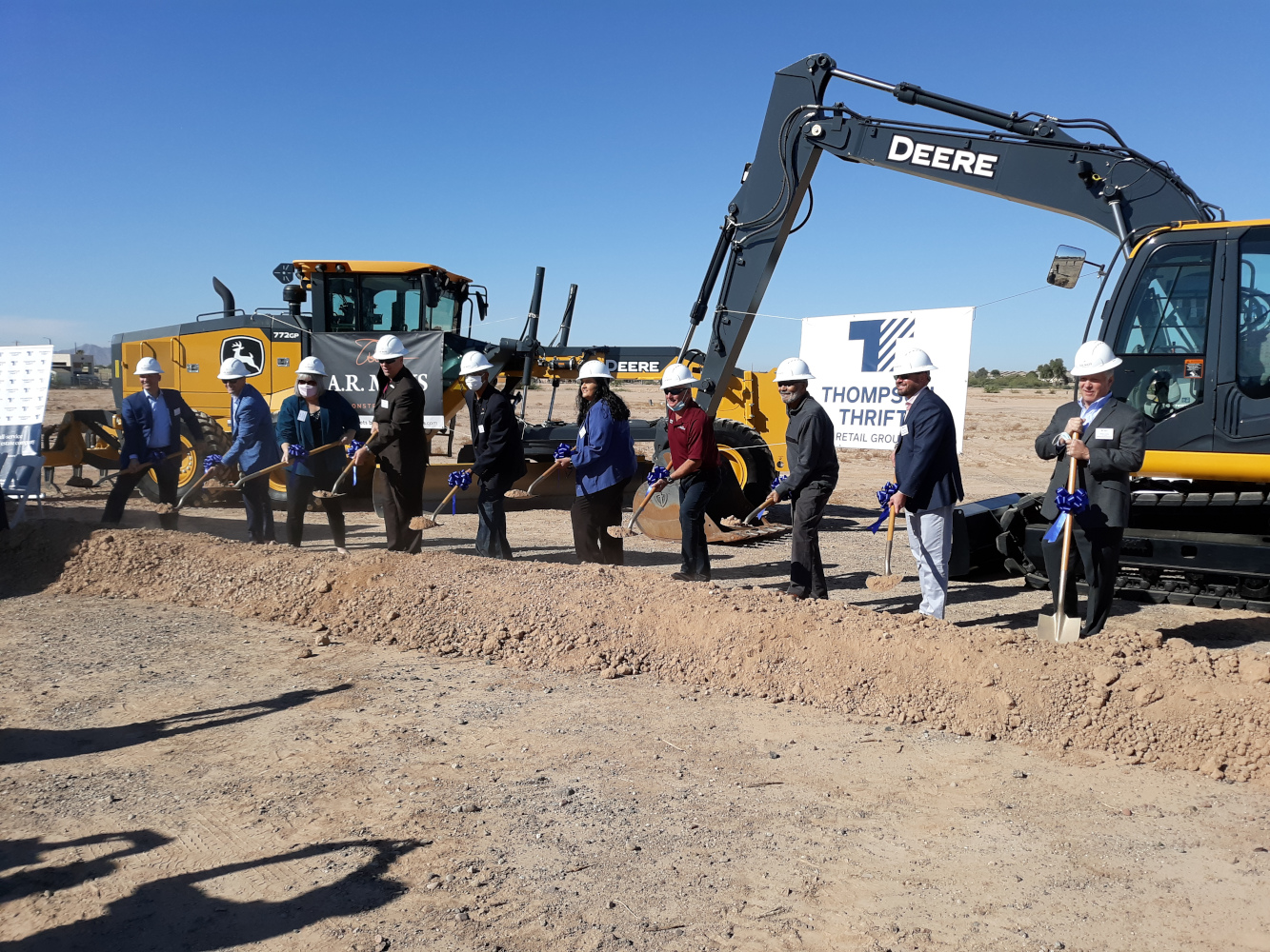 A group of people in hard hats and suits participate in a groundbreaking ceremony, each holding a shovel. Construction machinery and a 