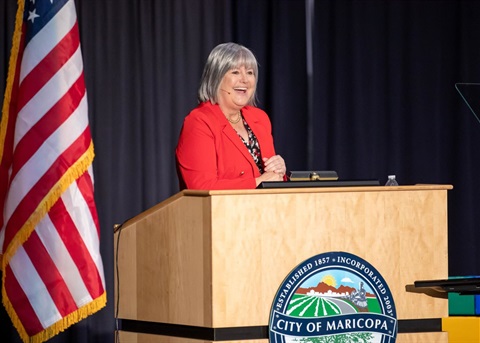 A woman in a red blazer speaks at a podium with the City of Maricopa seal. An American flag is visible. The atmosphere is professional and engaging.