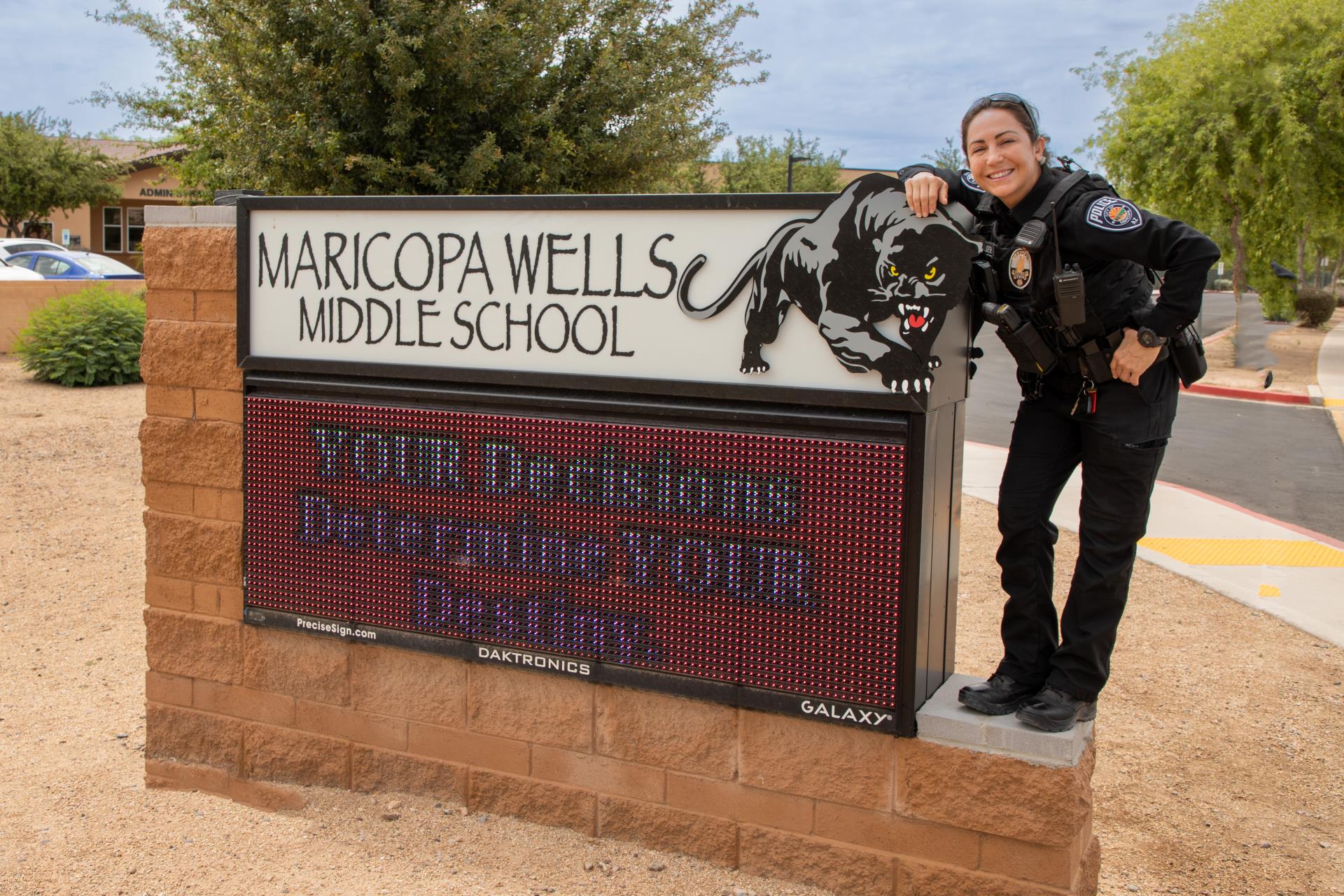 A smiling police officer stands beside the Maricopa Wells Middle School sign featuring a panther logo and the message, 