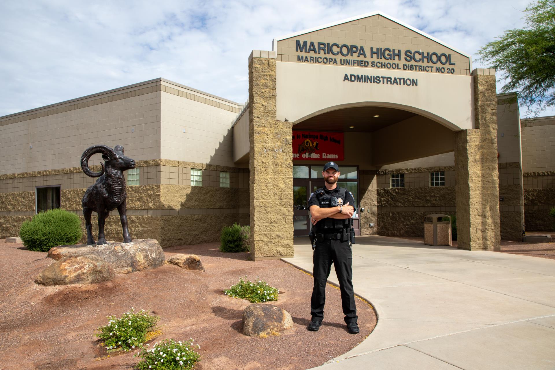 Security officer stands in front of Maricopa High School entrance, adjacent to a ram statue. Clear blue sky, conveying a sense of safety and vigilance.