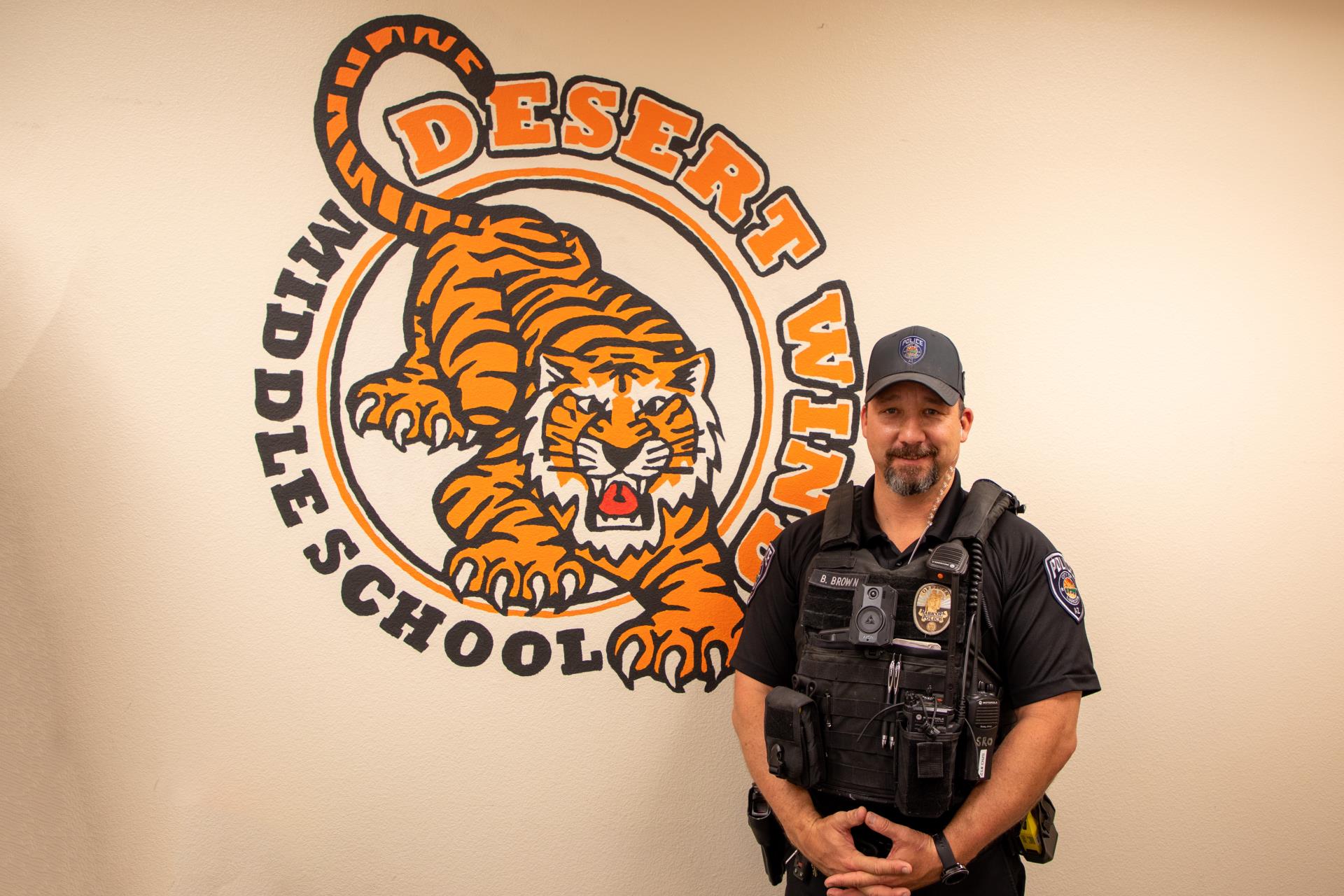 A police officer in uniform stands in front of a Desert Wind Middle School logo featuring a roaring tiger. The ambiance is professional and welcoming.