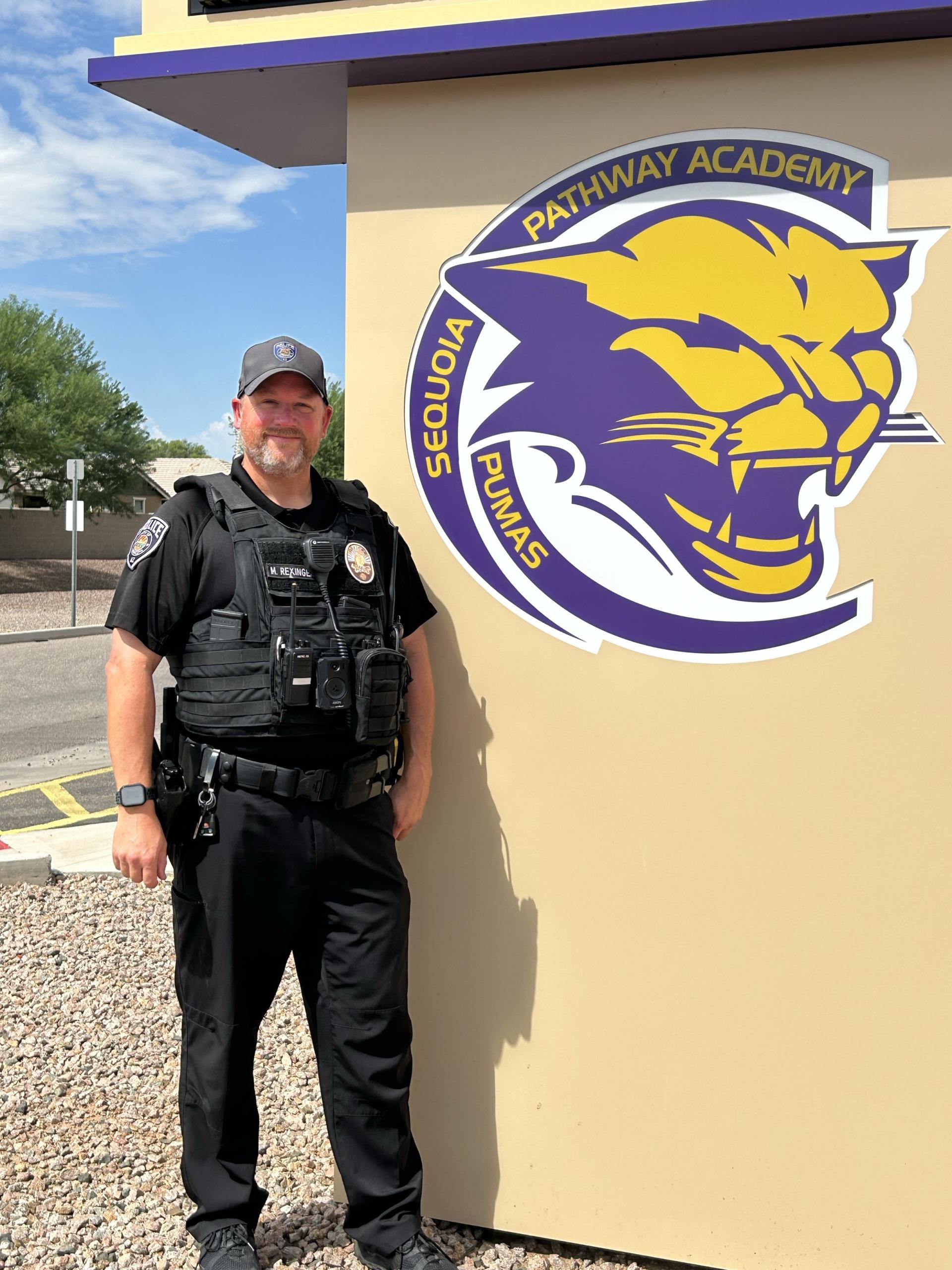 Security officer in uniform stands smiling beside a school sign featuring a fierce purple and yellow puma mascot, with clear blue sky in background.