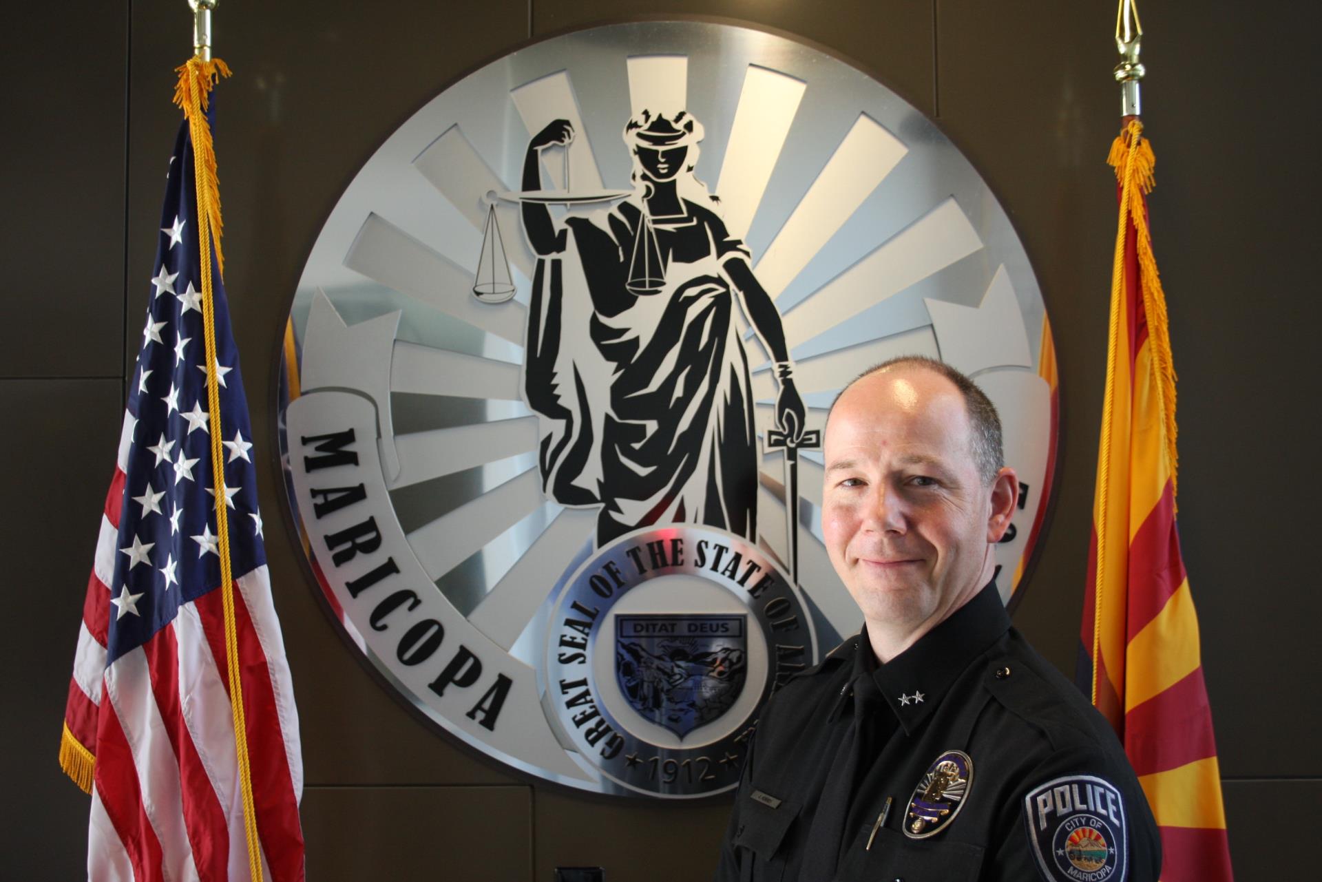 A police officer in uniform stands smiling in front of a circular police department emblem featuring a depiction of Lady Justice and a state seal.