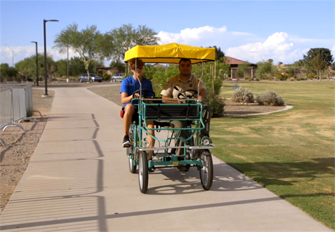 Two people ride a four-wheeled pedal cart with a yellow canopy on a sunny park pathway. The backdrop includes green grass, trees, and blue sky.