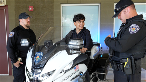 A young person sits on a police motorcycle, looking cheerful, flanked by two smiling police officers. The scene conveys a friendly and engaging interaction.