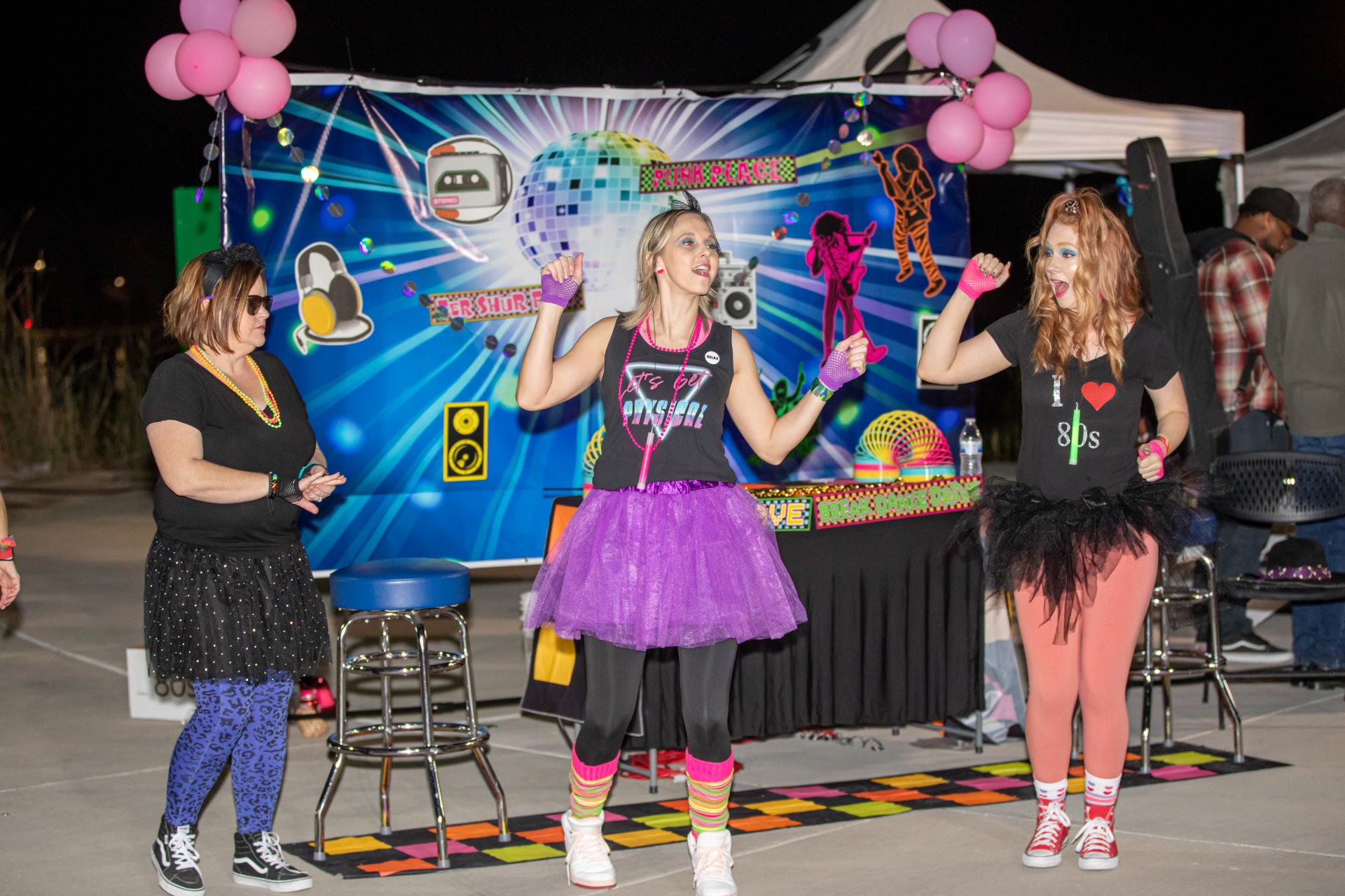 Three women dance at an 80s-themed party. They wear colorful tutus and leggings. A vibrant backdrop with a disco design adds to the festive mood.