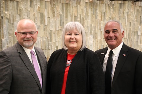 Three people standing together, smiling. They are dressed in formal attire with a stone wall backdrop. The mood is professional and cheerful.