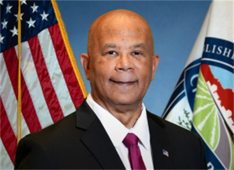 A man in a suit and tie stands before U.S. and organizational flags. He looks directly at the camera, conveying a formal and confident demeanor.