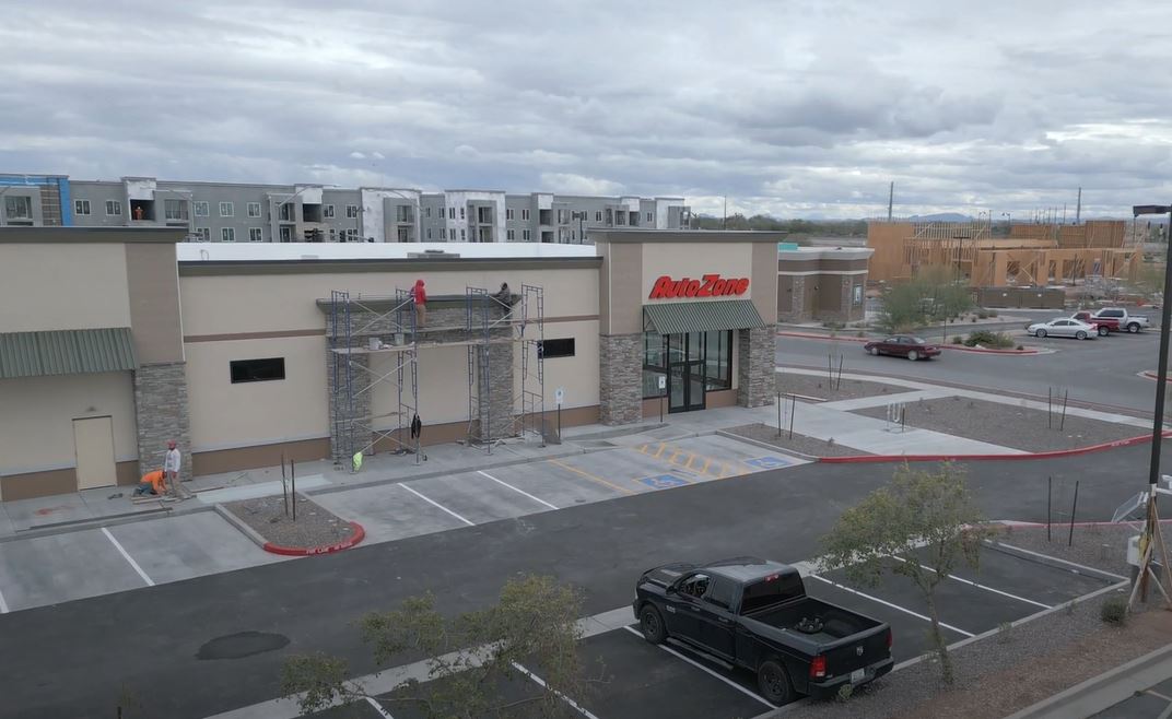 Aerial view of an AutoZone store under cloudy skies. A worker on scaffolding adjusts signage. An empty parking lot and scattered vegetation are visible.