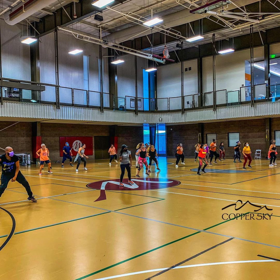 A vibrant group exercise class in a spacious gym. Participants enthusiastically follow a routine on a polished floor under bright overhead lights.
