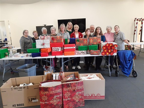 A group of smiling people stands behind a table filled with wrapped holiday gift boxes in various colors. The setting is festive and cheerful.