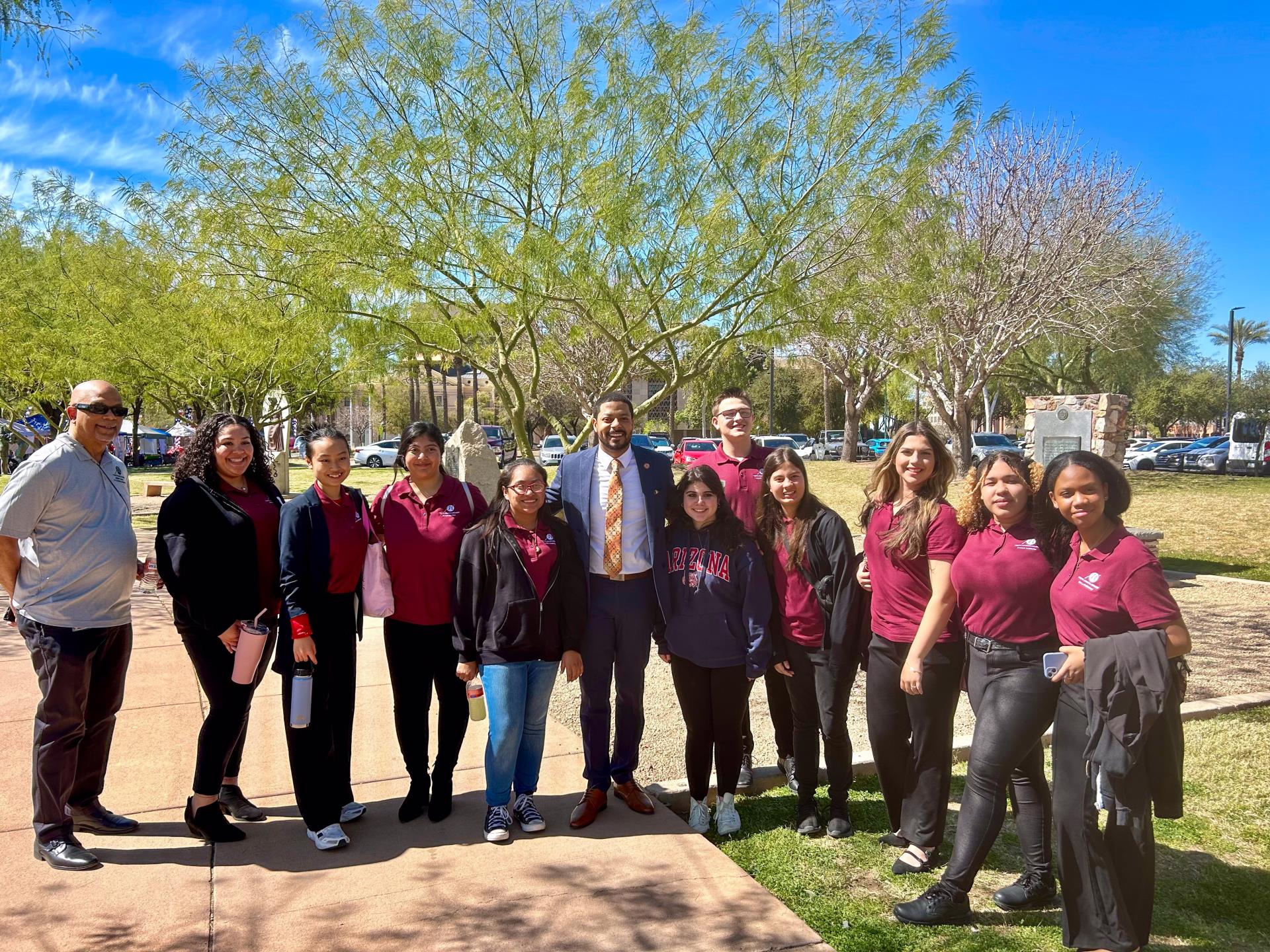 A group of people, some in maroon shirts, stand smiling outdoors on a sunny day. Trees with green leaves serve as a backdrop, conveying a cheerful mood.