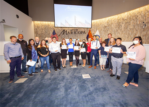 A diverse group of people stands smiling in a room, holding certificates. Behind them are U.S. and Arizona flags. The mood is celebratory.
