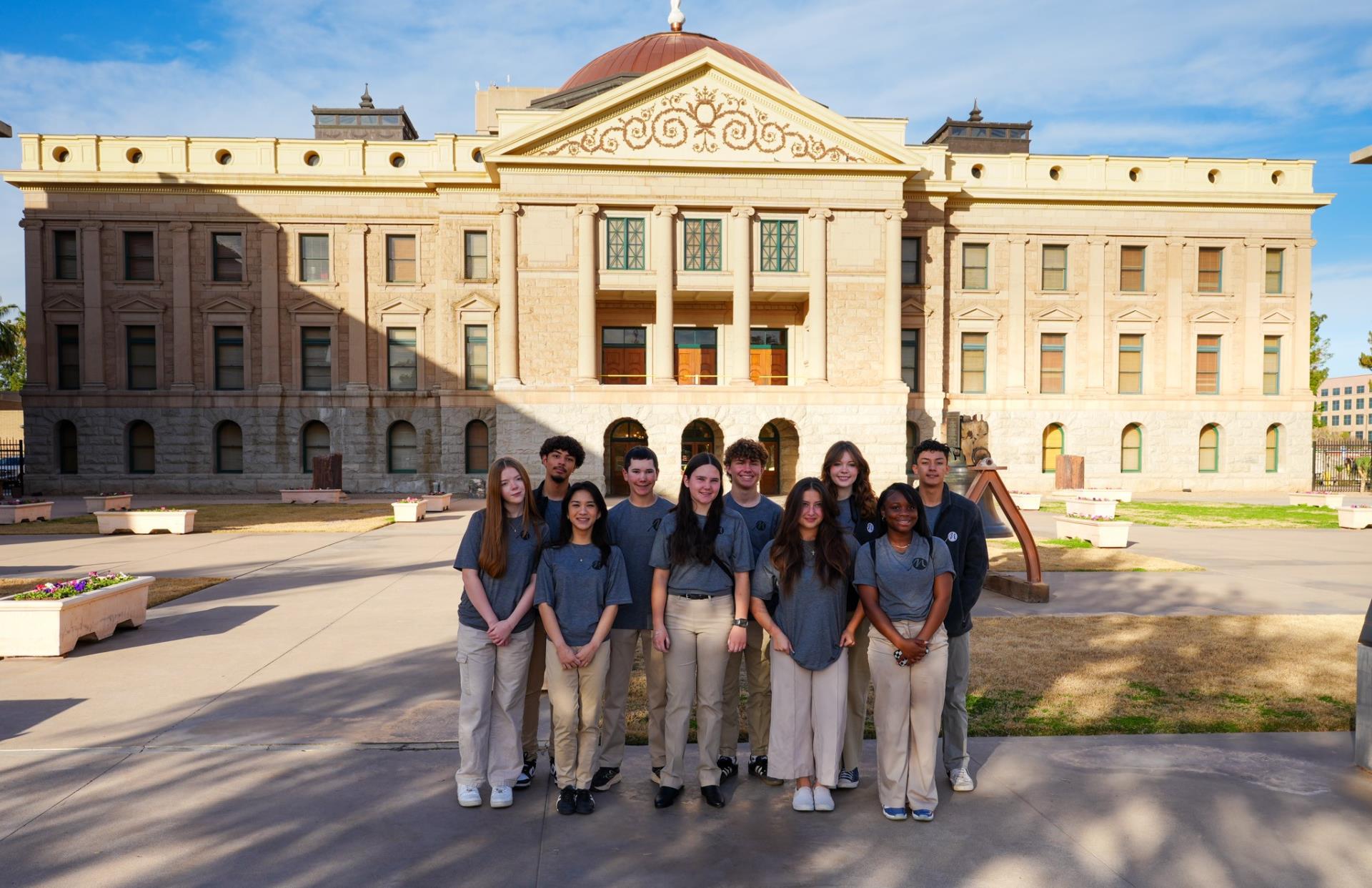 A group of young people in matching gray shirts and khaki pants stand smiling in front of an ornate, historic building under a clear blue sky.
