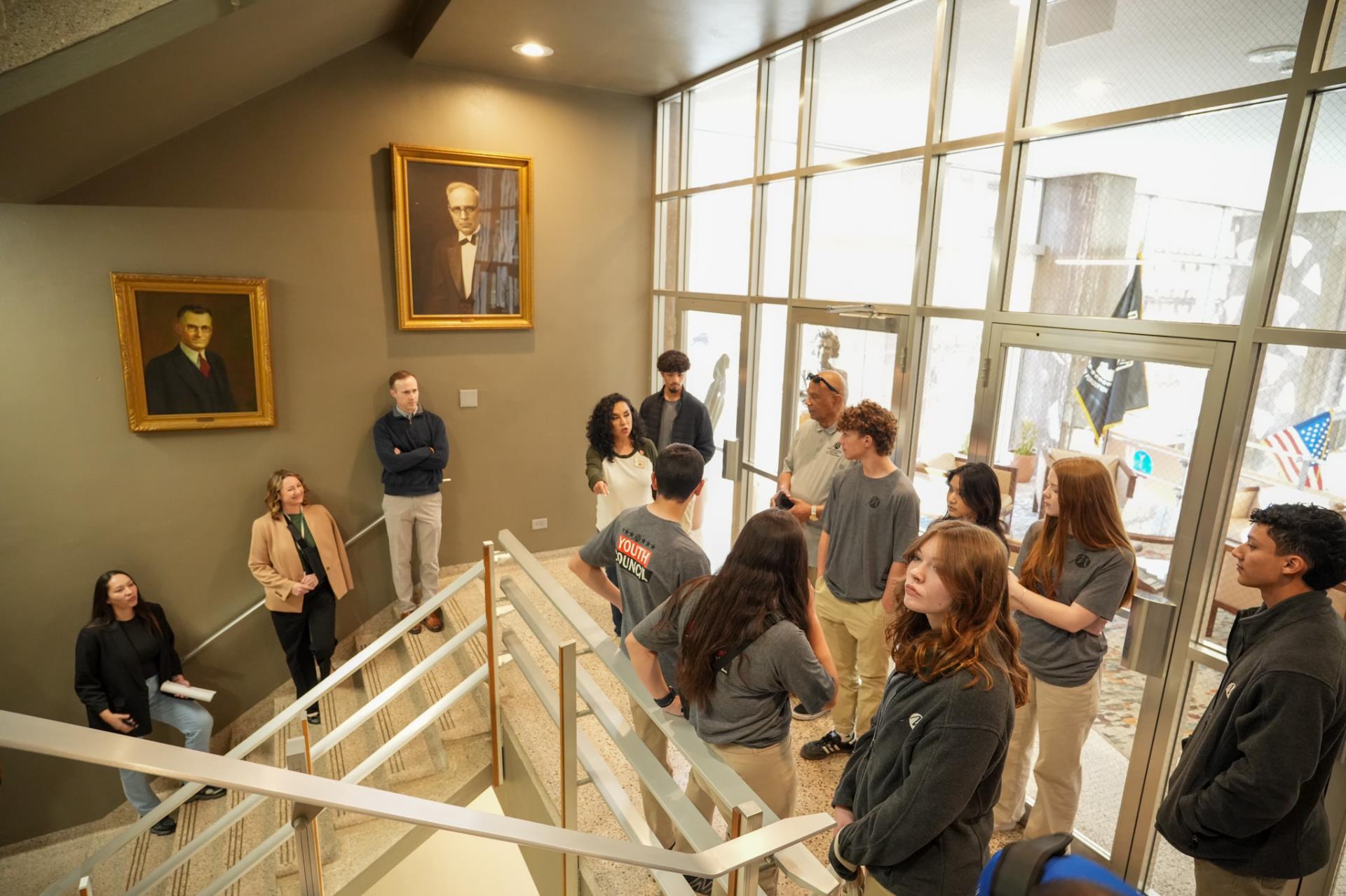 A group of students stand attentively on a stairway inside a building with large windows. Two portraits hang on the wall. The atmosphere is educational.