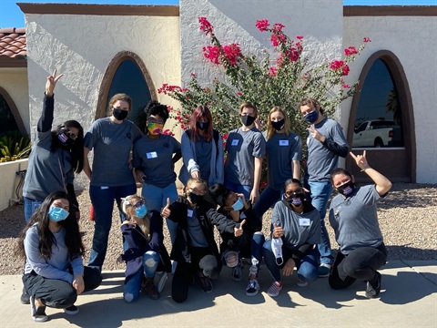 A diverse group of people wearing masks and matching gray shirts pose cheerfully, flashing peace signs and thumbs up, in front of flowering bushes and a stucco building.