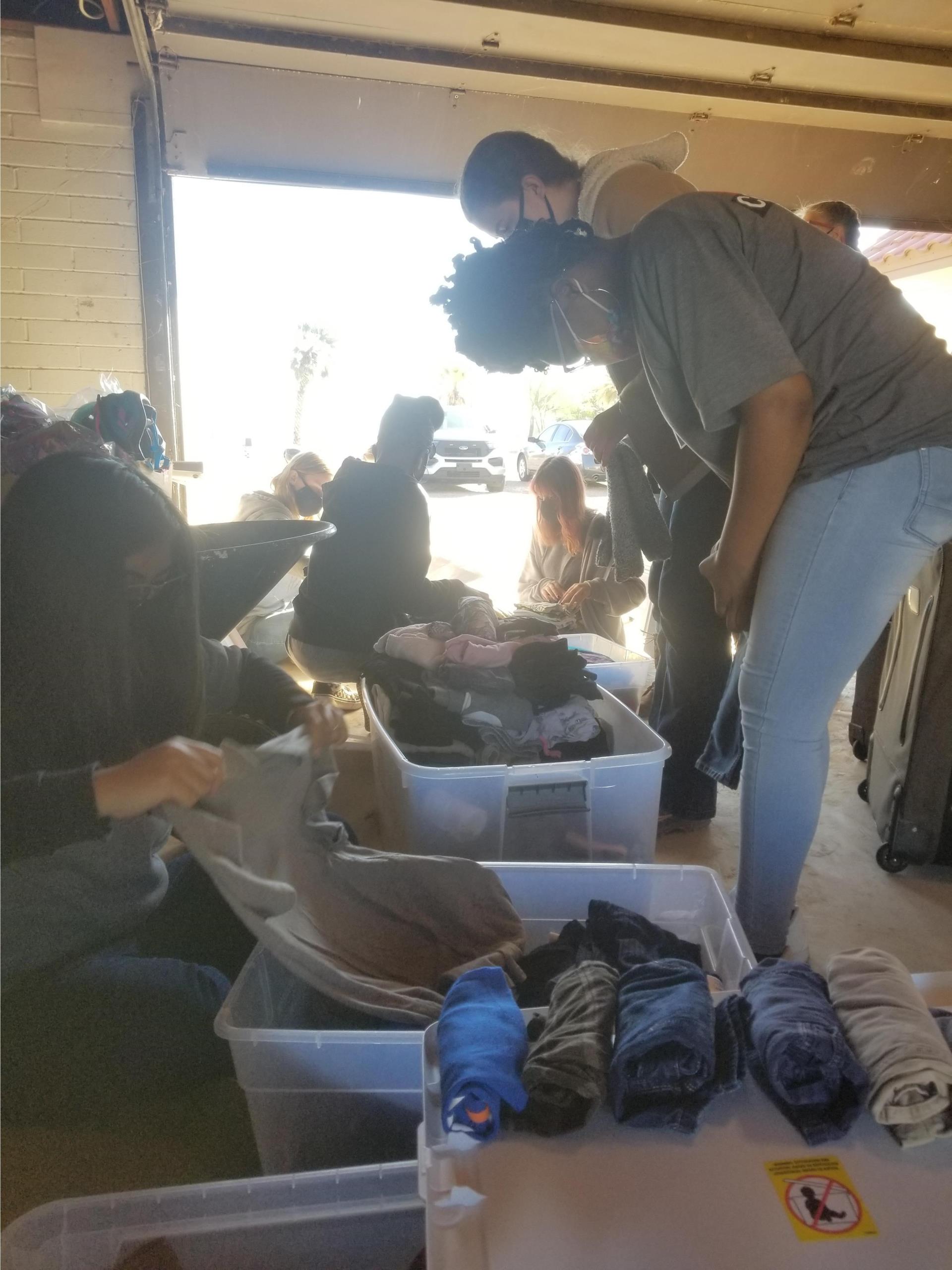 Group of people in a garage sort through bins of clothes, focusing intently. They wear casual attire and masks, conveying teamwork and community aid.