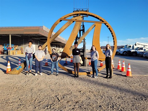 Six people stand in front of a large rusty metal sculpture amid a construction site. Orange cones and industrial vehicles are visible under a clear blue sky.