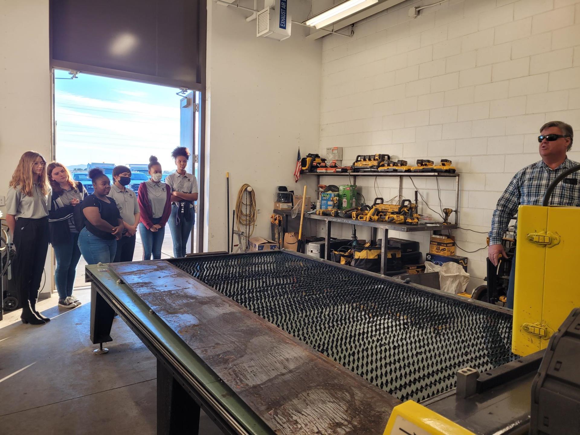 A group of people stands attentively in a workshop near a man speaking by a large industrial machine. The space has tools and equipment lining the shelves.