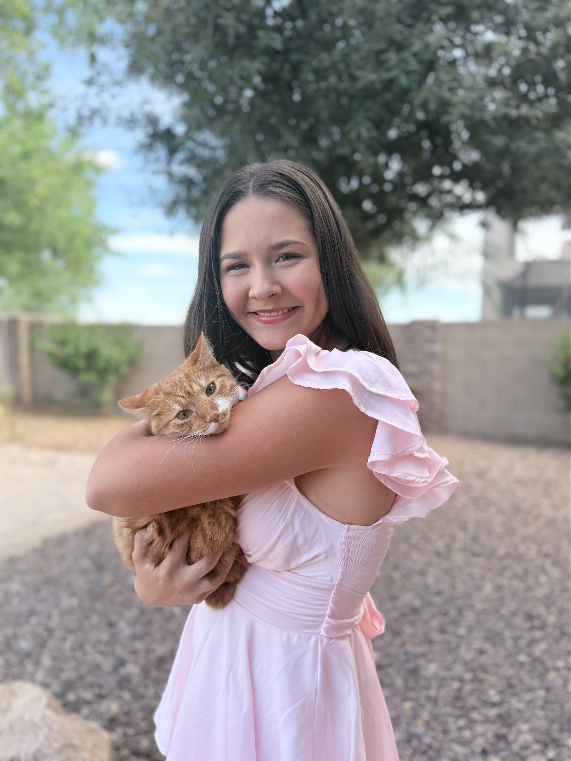 A woman in a pink dress smiles while holding an orange cat in an outdoor setting. There are trees and a stone wall in the background, creating a warm, joyful atmosphere.