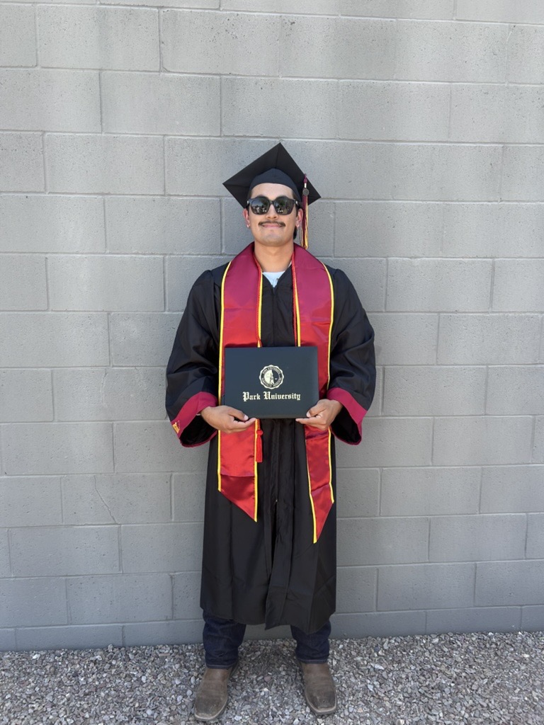 A graduate wearing a black cap and gown with red and yellow trim holds a Park University diploma. He stands confidently against a gray brick wall.