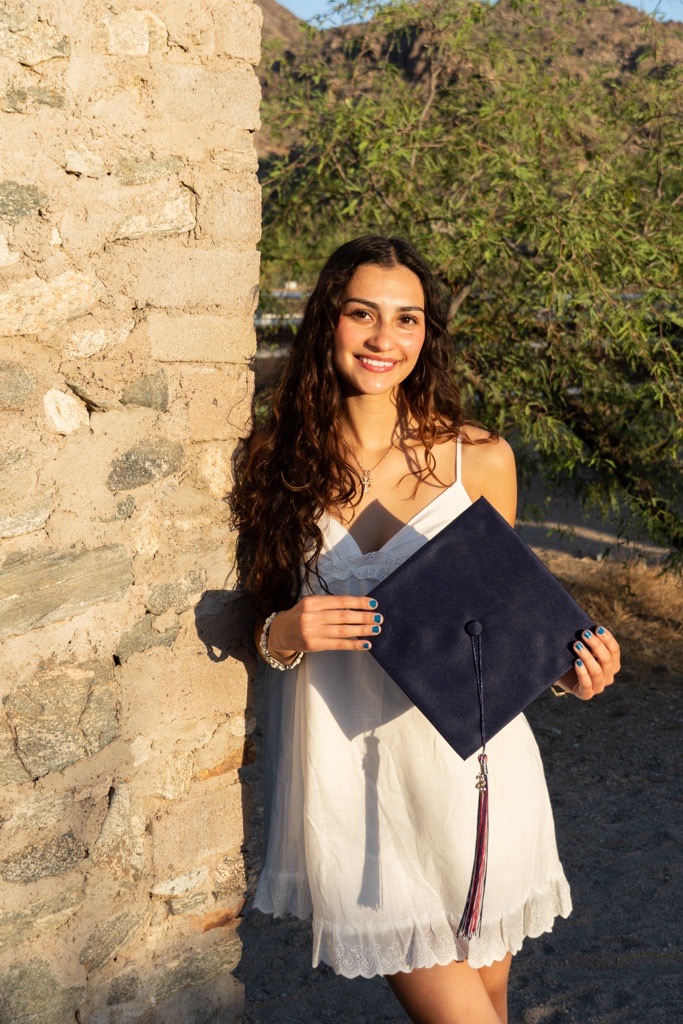 Smiling woman in a white dress holds a navy graduation cap by a stone wall. Sunlight and nature in the background create a joyful, celebratory tone.