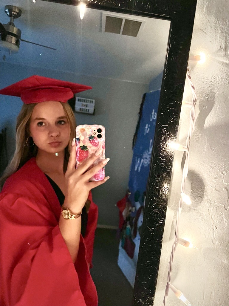 Young woman in a red graduation gown and cap takes a mirror selfie. Room is softly lit with string lights, creating a celebratory atmosphere.