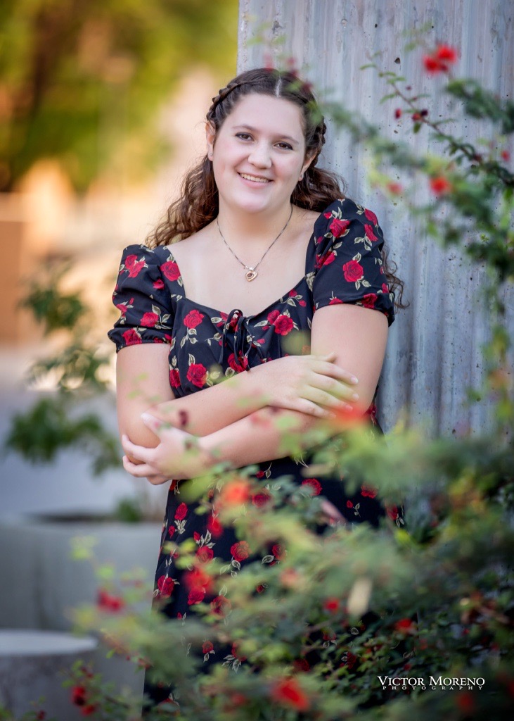 Young woman with braided hair in a floral black dress leans against a concrete wall, smiling gently. Surrounded by greenery and red flowers.