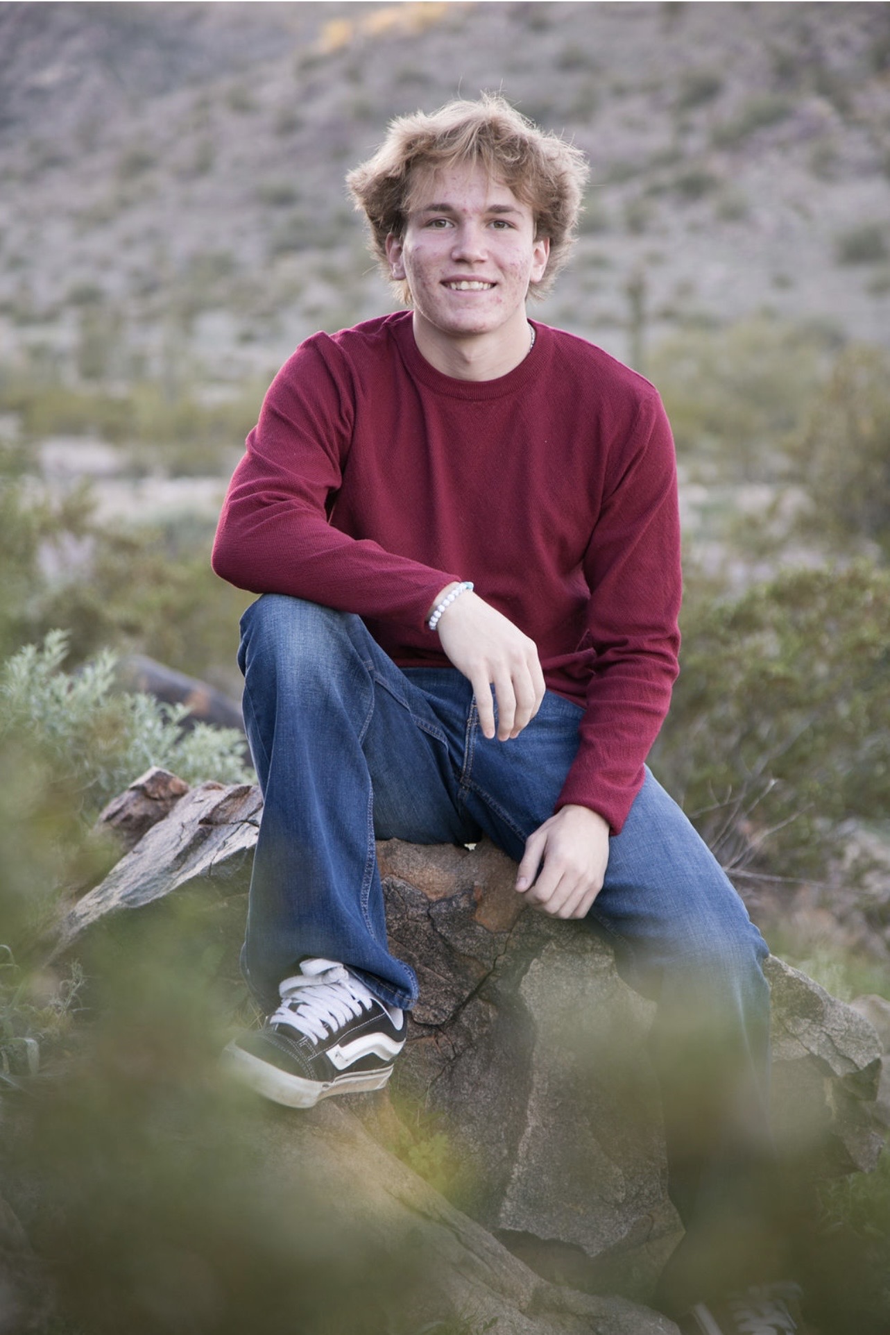A young man in a red sweater and jeans sits on a rock, smiling. He is outdoors with a blurred desert landscape in the background, conveying a relaxed, natural vibe.