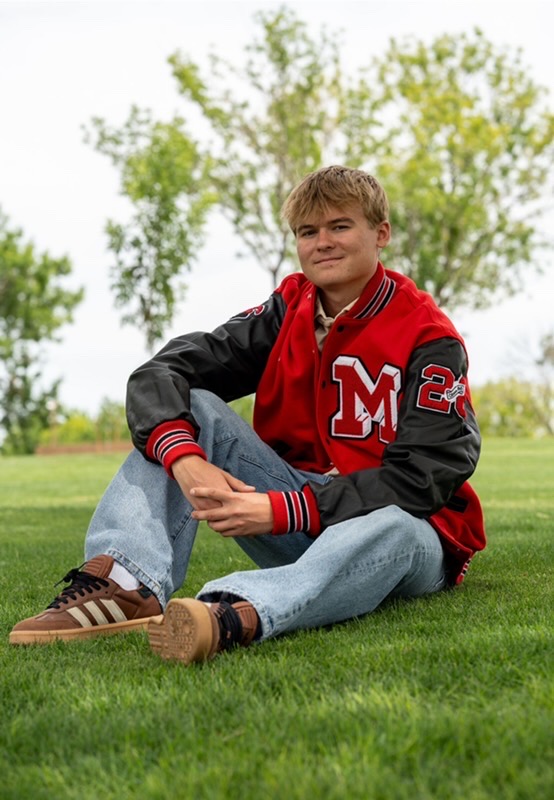 Young man sitting on grass in a red varsity jacket with a large 