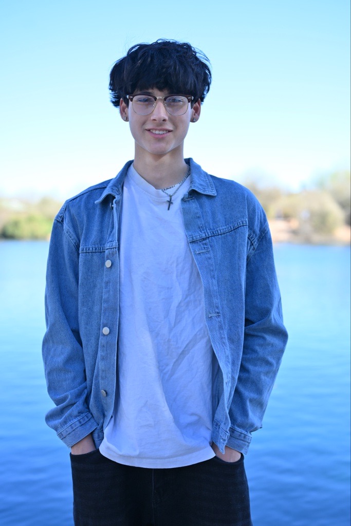 Young person with glasses smiling, wearing a denim jacket and white shirt. They stand near a lake, with a clear blue sky in the background.