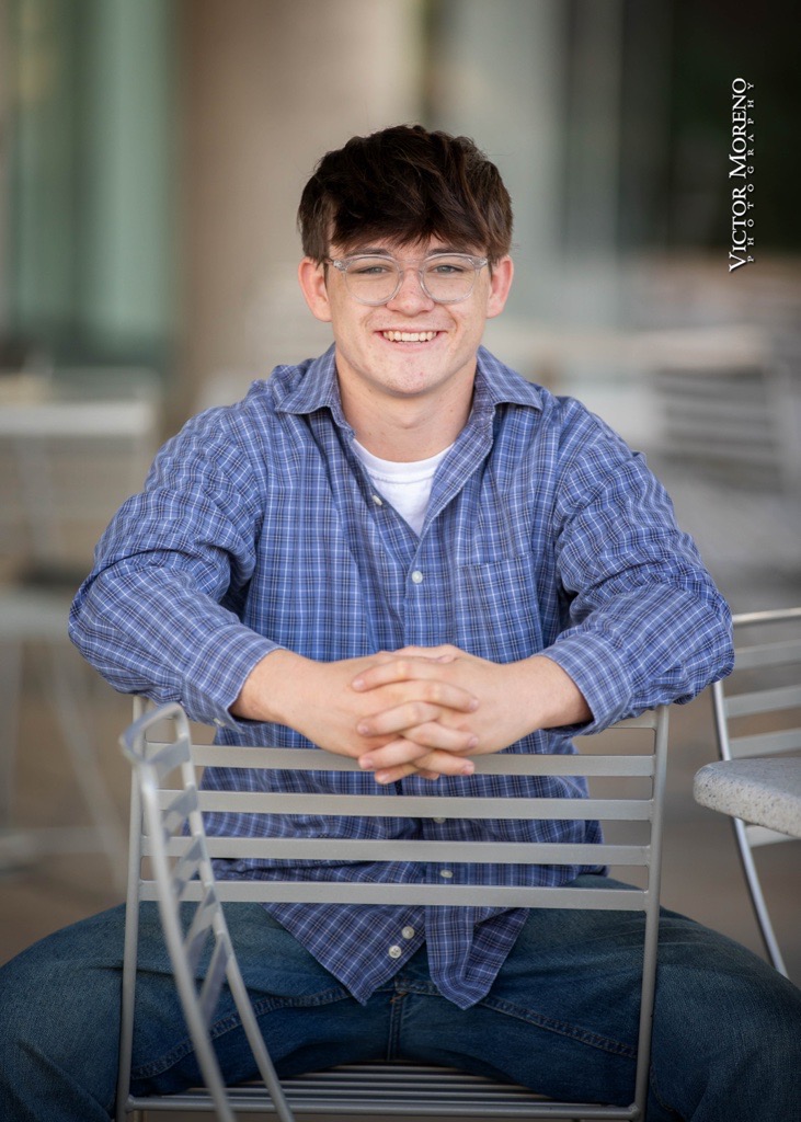 Young man with short hair and glasses sits on a metal chair backward, wearing a blue plaid shirt and jeans. He smiles warmly in a casual, outdoor setting.