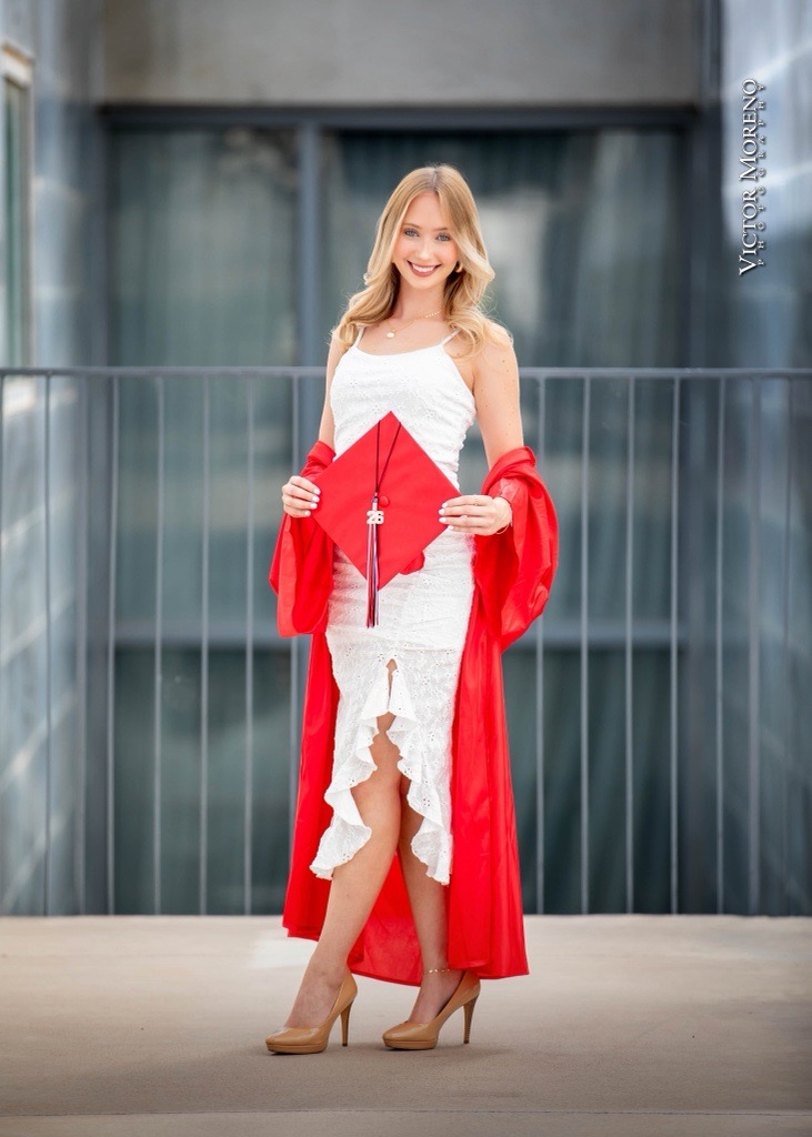 Woman in a white lace dress and red graduation gown holding a cap stands smiling. Background features a blurred modern building facade.