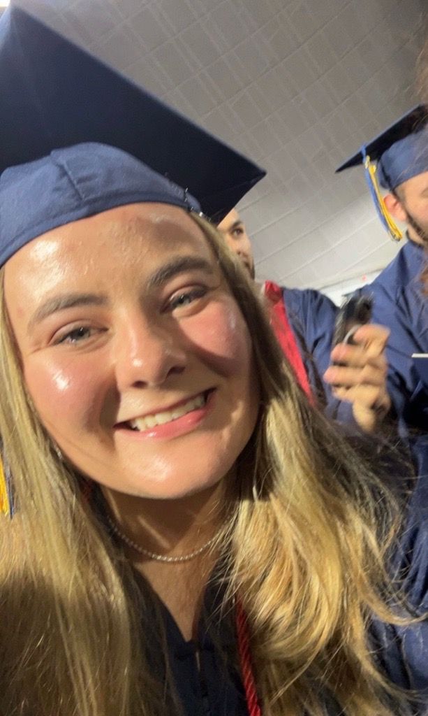 Smiling graduate in a blue cap and gown takes a selfie indoors. Her expression conveys joy and achievement. Others in similar attire are in the background.