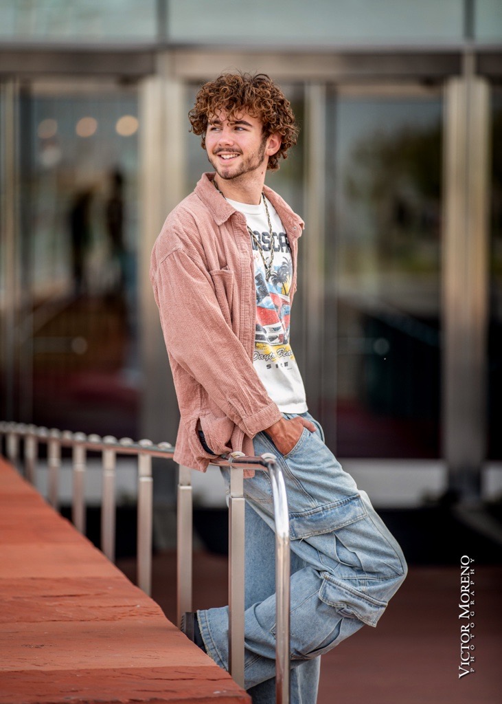 Young man with curly hair leans casually on a railing, wearing a pink jacket and jeans, smiling, with a blurred modern building entrance behind him.