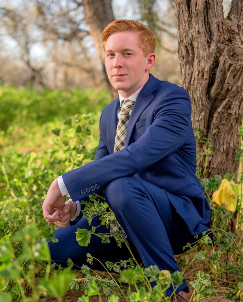 Young man with red hair in a blue suit and plaid tie crouching beside a tree in a green, sunlit forest. He appears thoughtful and confident.
