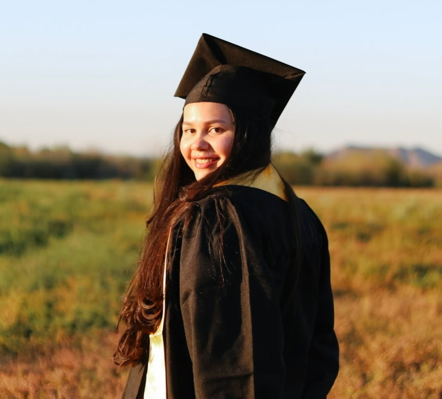 A smiling graduate in a black gown and cap stands in a sunny field, looking over her shoulder. The scene conveys a sense of achievement and joy.