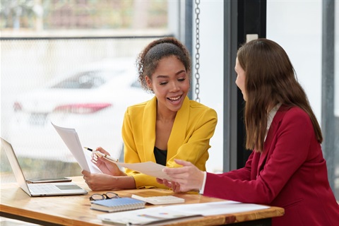Two women in blazers engage in a discussion at a wooden table, reviewing papers in a bright, well-lit setting.