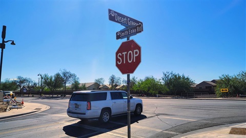 Street intersection with a STOP sign, “Desert Greene” and “Smith Enke Rd” street signs, a white SUV, desert homes, and construction barricades under a clear blue sky.