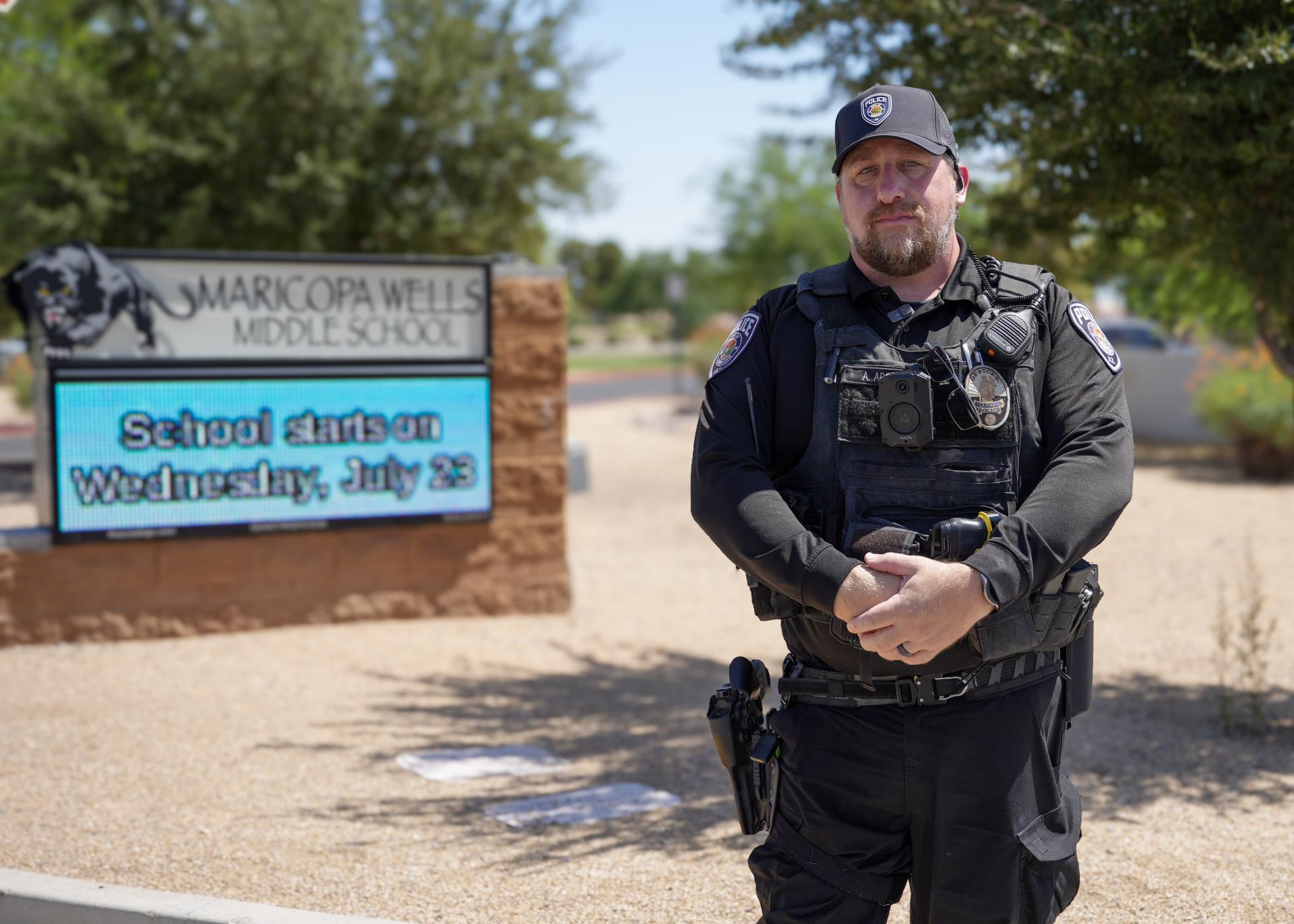Police officer in uniform stands in front of Maricopa Wells Middle School entrance sign. The sign shows a message about the school start date.