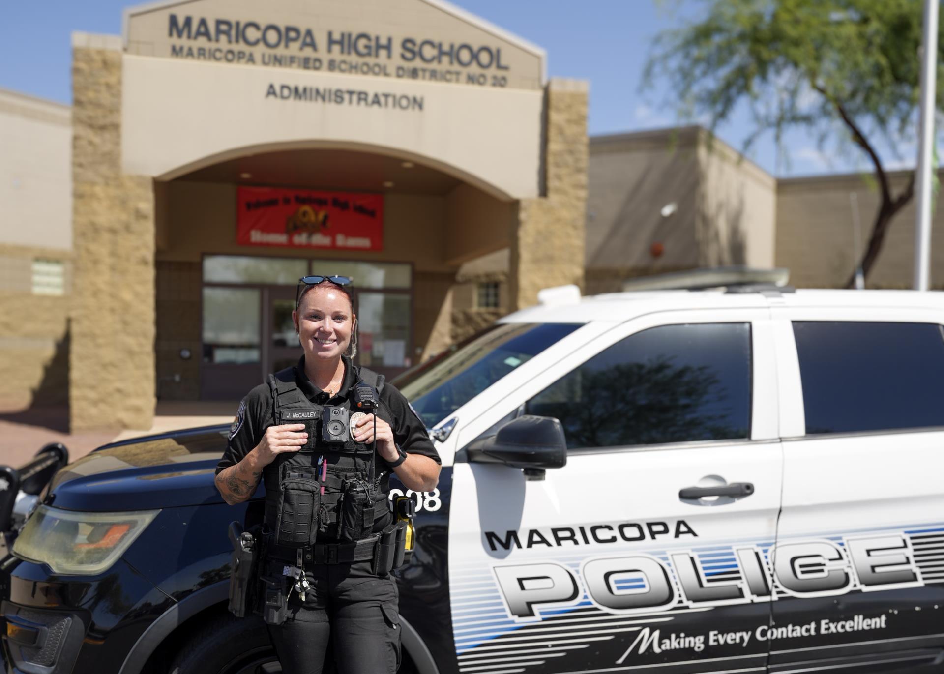 A police officer stands smiling in front of a Maricopa Police car, parked outside Maricopa High School's entrance, conveying a sense of safety and community.