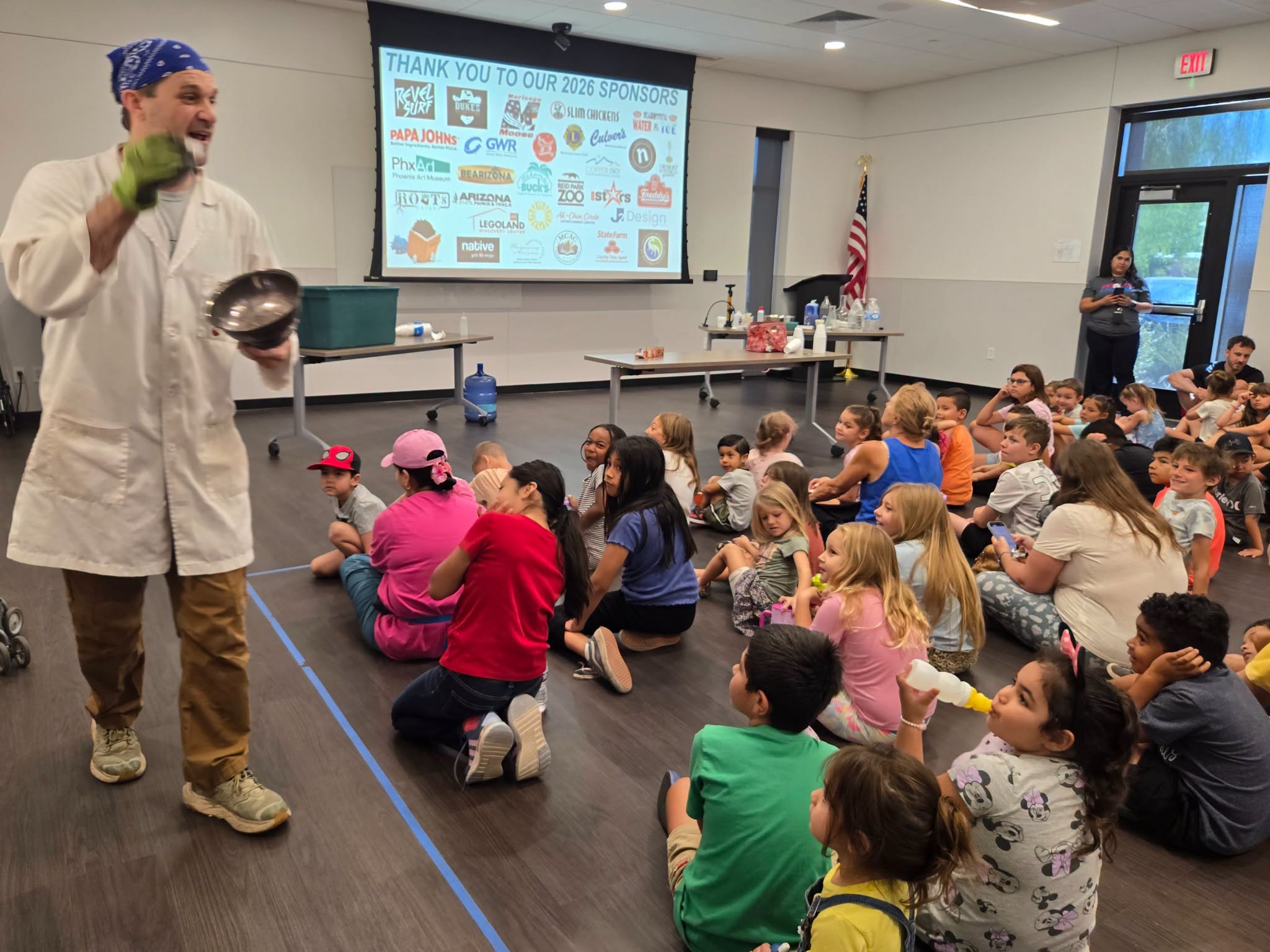 A male presenter in a white coat and blue bandana gestures with a metallic bowl in front of an audience of children sitting on the floor in a modern classroom.